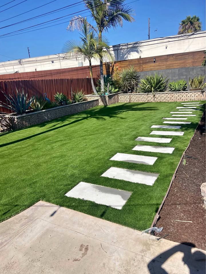 A backyard with bright green grass installed by an expert artificial turf installer in Queen Creek, large square stepping stones forming a path, palm trees, and lush plants along the fence under a clear blue sky.