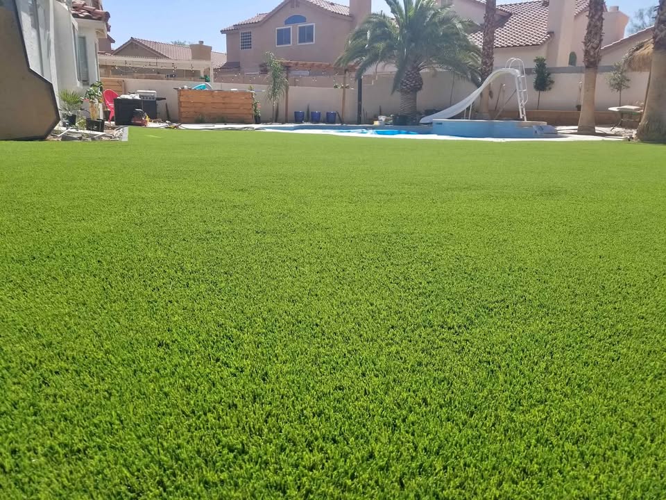 A backyard with bright green artificial grass installed by an expert artificial turf installer in Queen Creek, a swimming pool with a slide, palm trees, and several houses in the background under a clear blue sky.