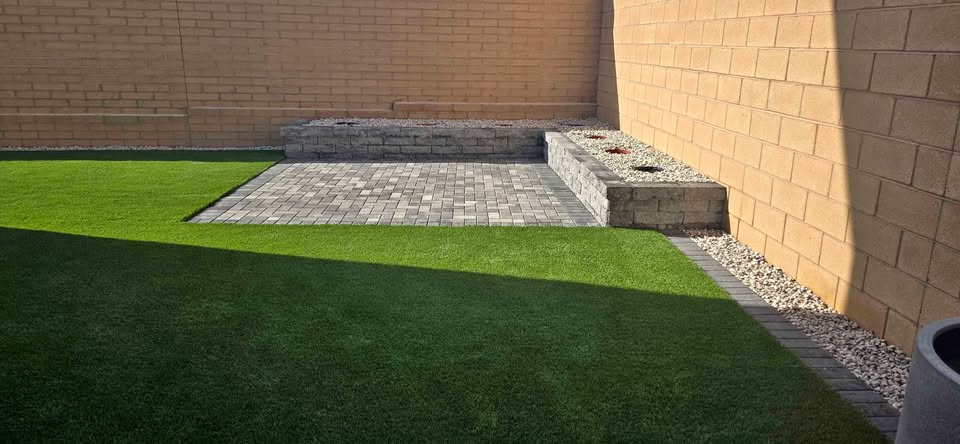 A backyard by Expert Artificial Turf Installer Queen Creek features artificial grass, a raised stone garden bed in the corner, gray stone pavers for the patio, and a brick wall. White decorative rocks accent the garden bed and edges.