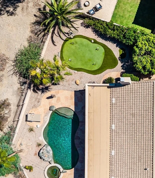 Aerial view of a backyard with a kidney-shaped pool, attached hot tub, lounge chairs, and a small putting green expertly crafted by an Expert Artificial Turf Installer Queen Creek, all surrounded by trees and landscaping.