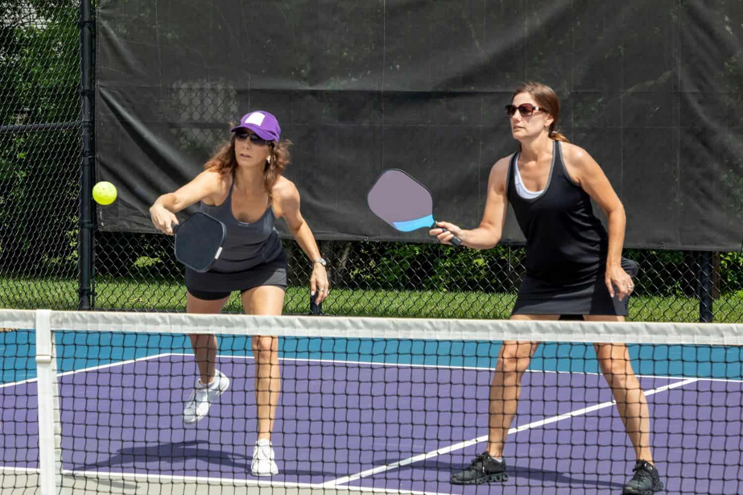 Two women playing pickleball on an outdoor court with sports field artificial turf. Both are focused on the ball, holding paddles, and wearing athletic clothing. One is about to hit the ball while the other stands ready. A black fence is in the background.