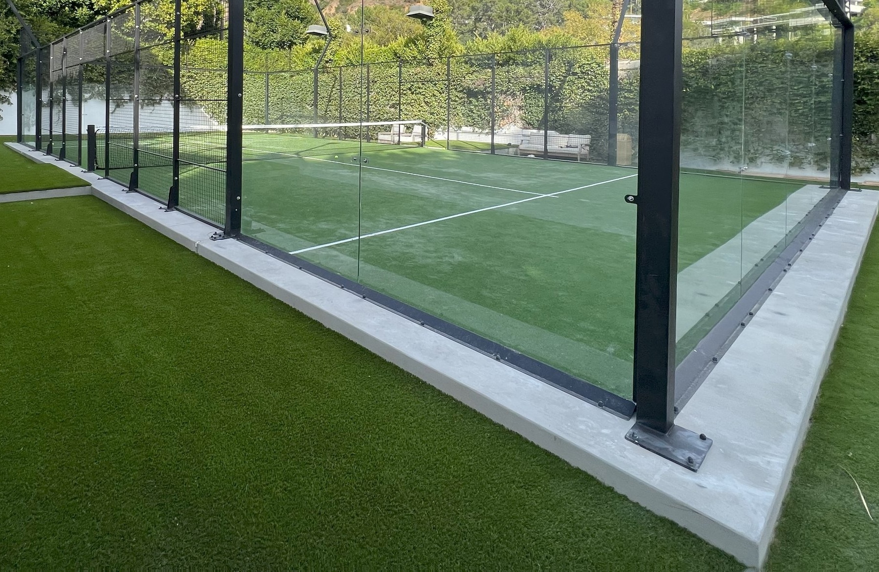 A padel tennis court surrounded by glass walls and metal posts, with sports field artificial turf outside the court and lush greenery visible in the background.