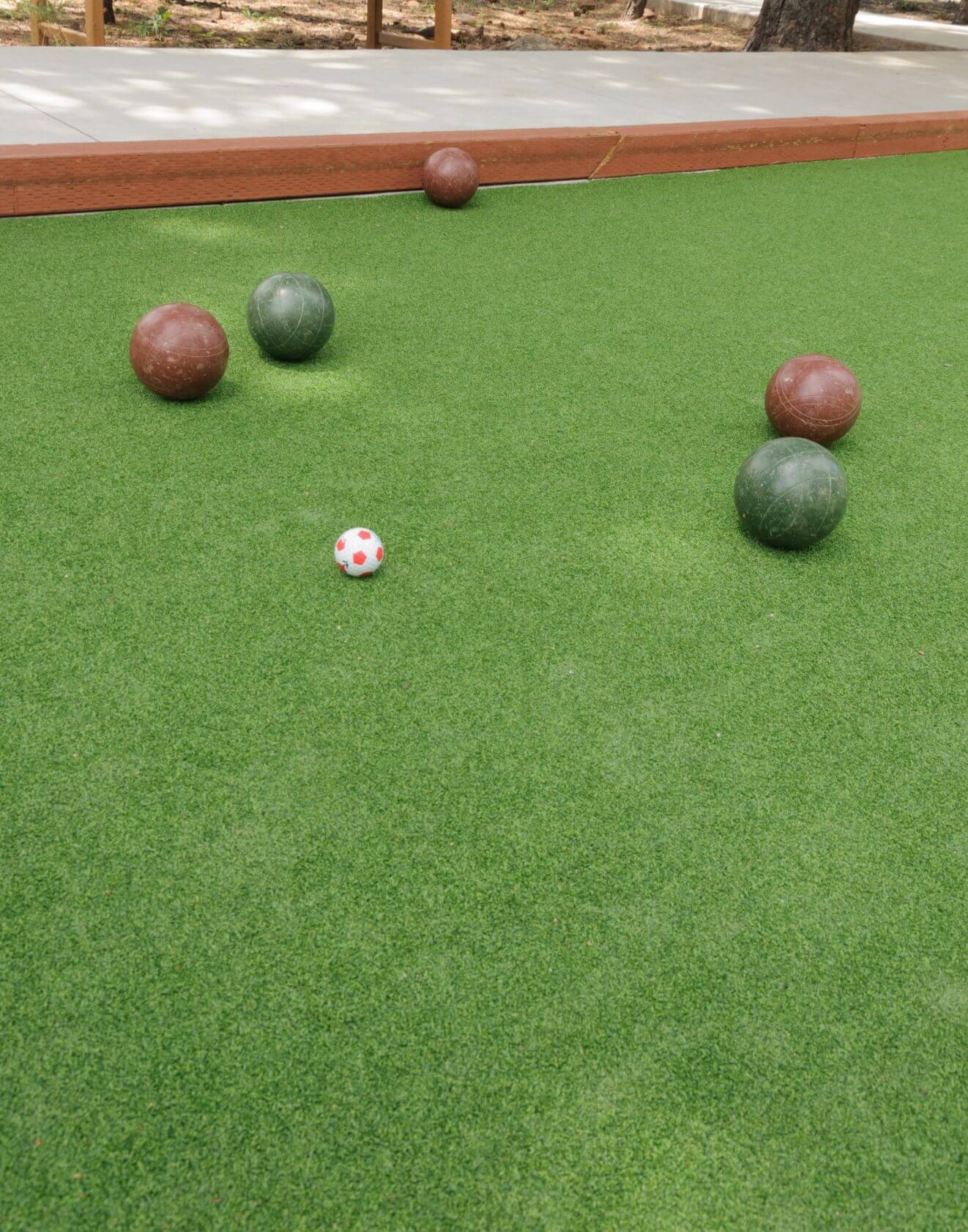 Several bocce balls in green and brown are arranged on a sports field with artificial turf, centered around a small white and red pallino ball. A wooden border surrounds the court.