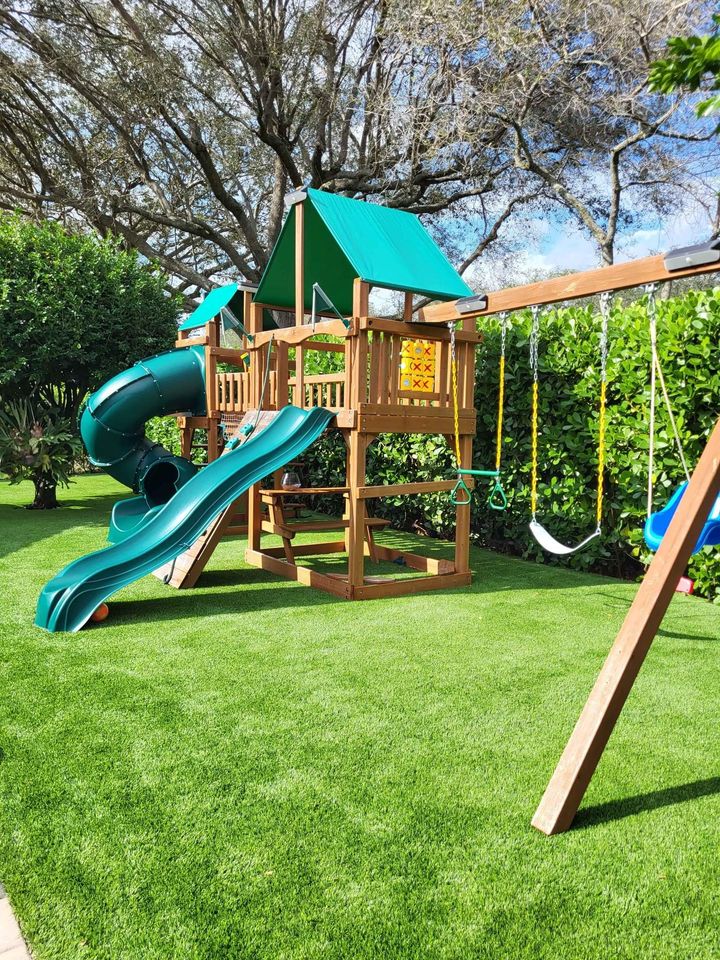 A wooden playground set on Playground Artificial Grass Queen Creek features slides, swings, a climbing wall, and a green canopy, surrounded by lush bushes and tall trees under a blue sky.