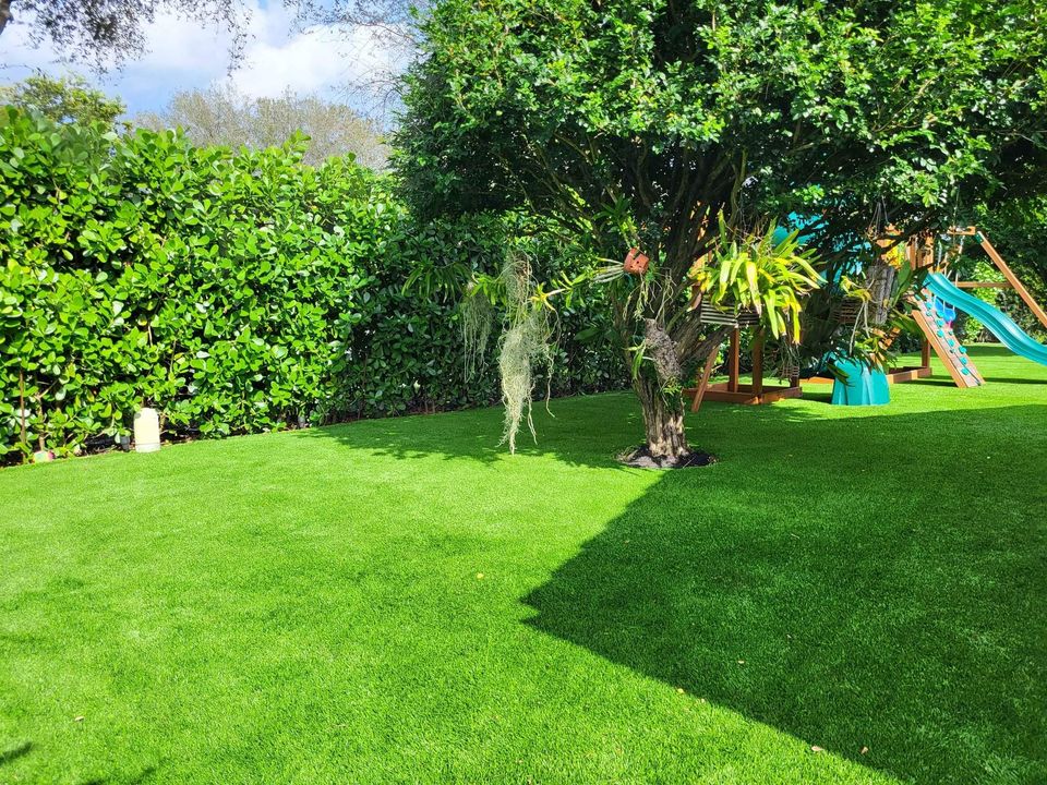 A lush green backyard with Playground Artificial Grass Queen Creek features a large leafy tree and a children's playset with slides. Sunlight creates shadows across the turf, while dense hedges line the yard for added privacy.