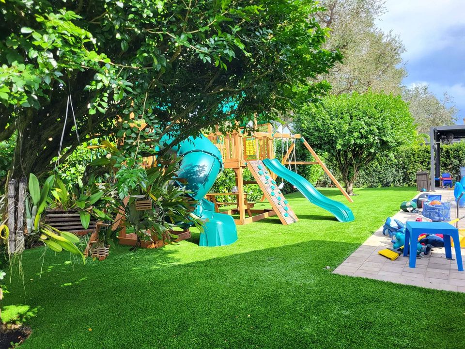 A backyard with lush Playground Artificial Grass Queen Creek, a large wooden playset featuring teal slides and a climbing wall, surrounded by trees and plants. Nearby are blue and red plastic chairs and toys on a paved area.