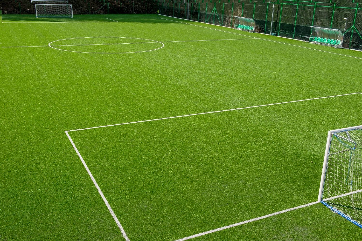An empty outdoor sports field features bright green artificial turf, crisp white boundary lines, two goals, and a row of green benches along one side, all enclosed by a tall fence.