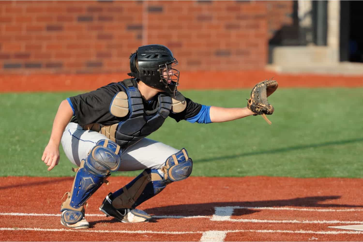 A baseball catcher in full gear crouches behind home plate on a sports field with artificial turf, wearing a helmet, chest protector, and shin guards, glove extended and ready to catch a pitch on a sunny day.