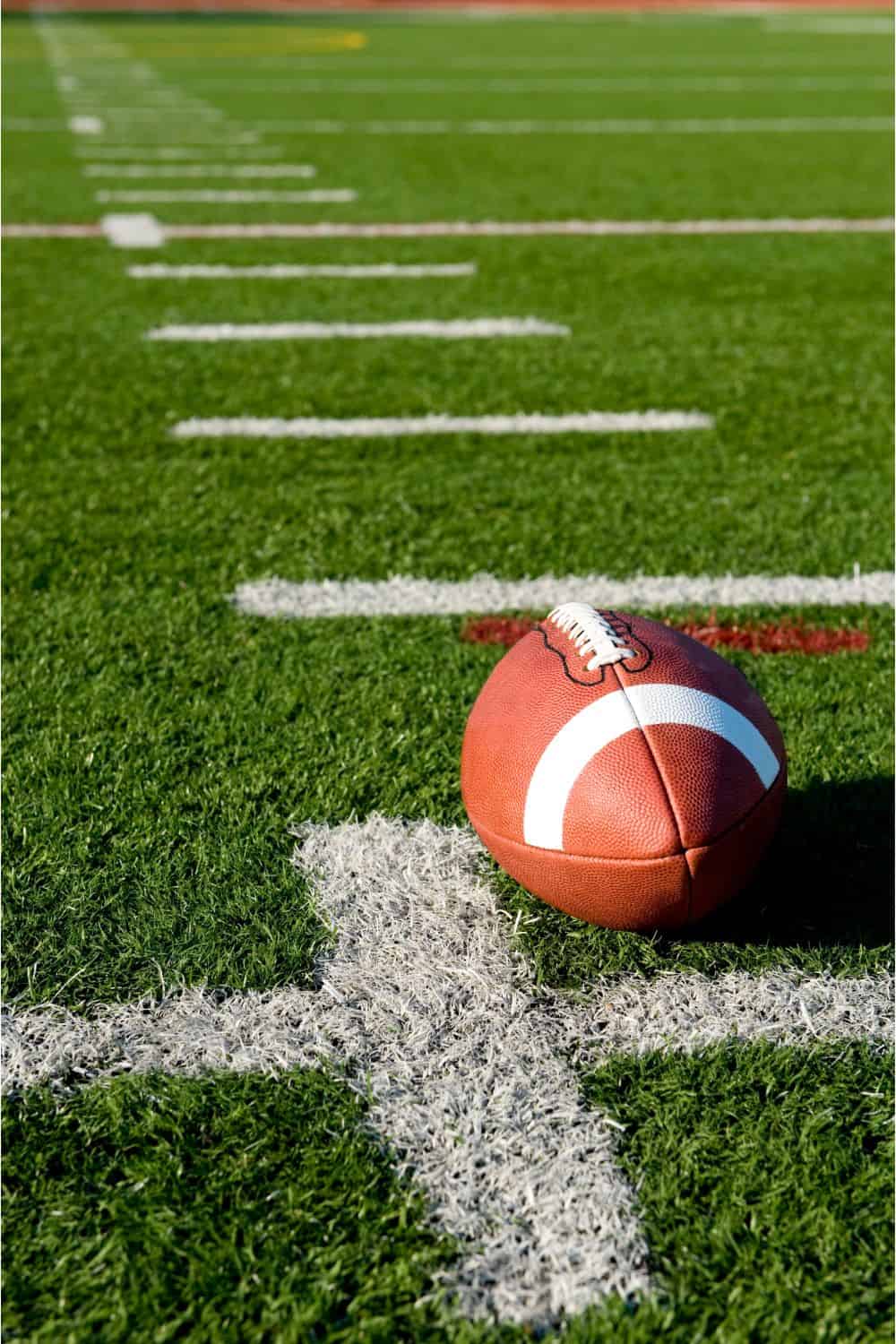 An American football rests on the green sports field of artificial turf near a white yard line, with more yard lines visible in the background under bright sunlight.