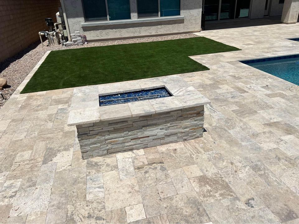 A modern backyard patio by Paver Installation Queen Creek features light stone tiles, a sleek rectangular fire pit, artificial grass, and a swimming pool beside a house with large windows.